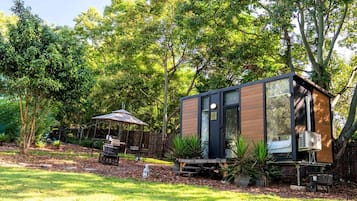A modern, compact outdoor structure with a door and windows, surrounded by lush greenery and a patio area with a table and chairs.
