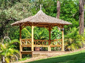 Un gazebo de madera con techo de paja, rodeado de abundante follaje y árboles.