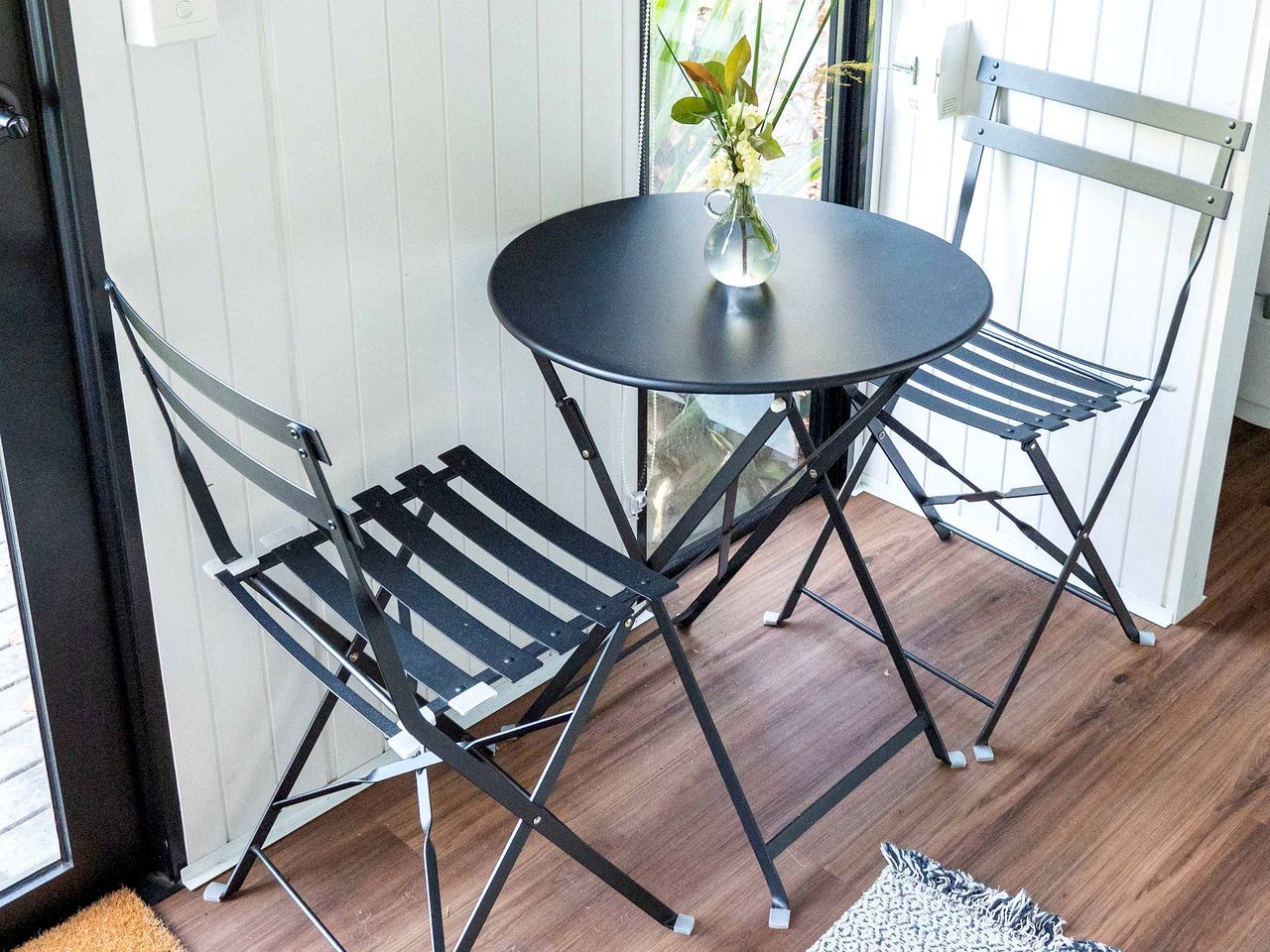 Une petite table ronde surmontée d’un vase de fleurs, deux chaises en métal noir et un plancher en bois.