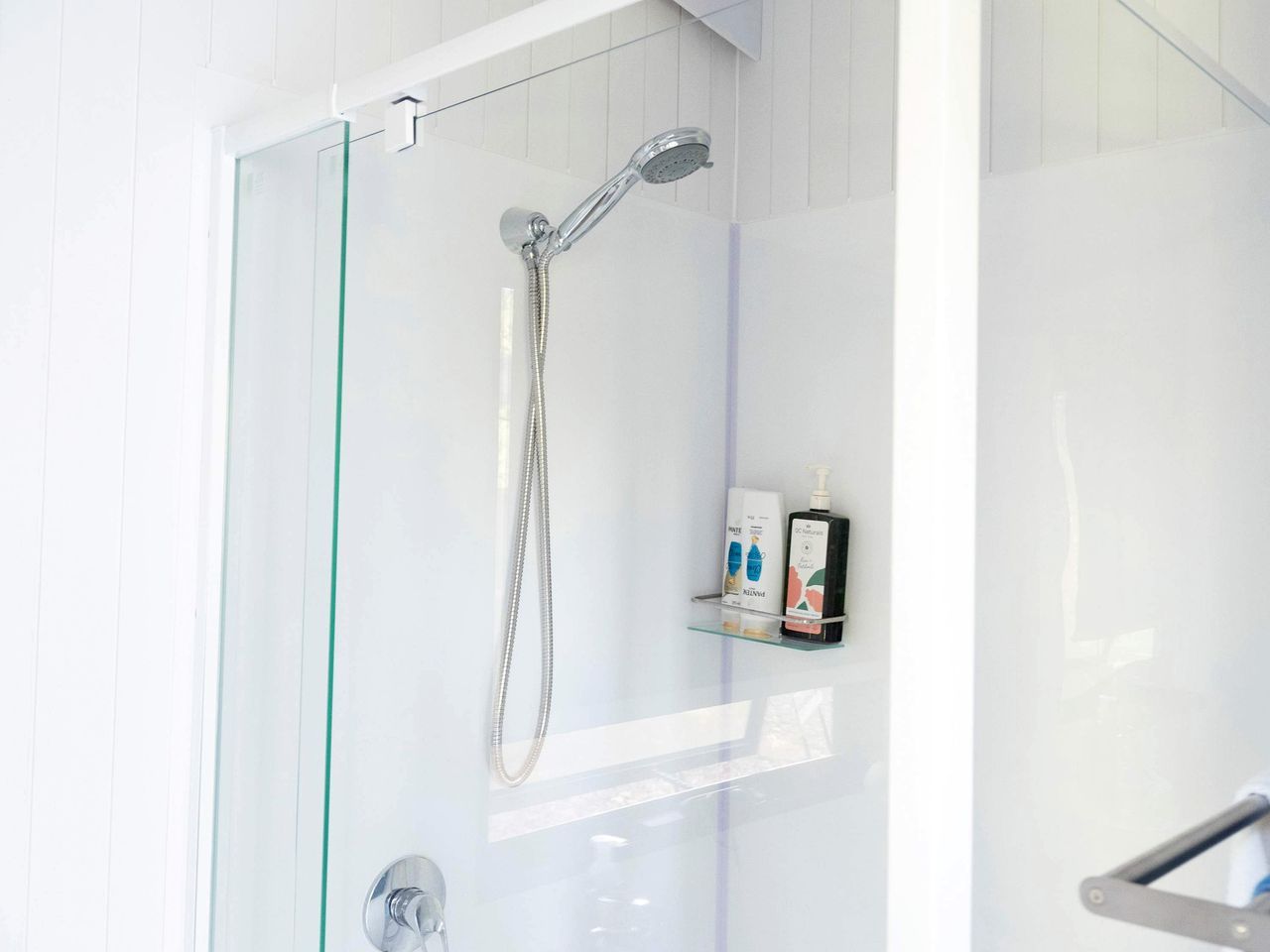 A glass-enclosed shower with a handheld showerhead and a glass shelf with toiletries.