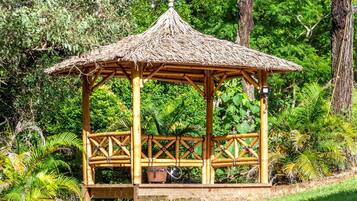 A wooden gazebo with a thatched roof, surrounded by lush greenery and trees.