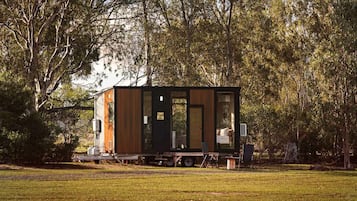 Une petite cabane moderne, dotée d’une grande porte coulissante, située dans une zone herbeuse, avec des arbres en arrière-plan.