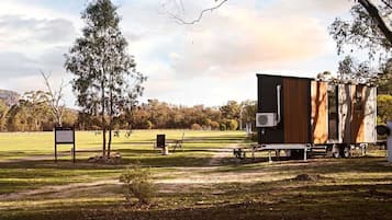 Een stacaravan met een groot raam en een klein bijgebouw, gelegen in een begrazend veld met bomen en een heldere lucht.