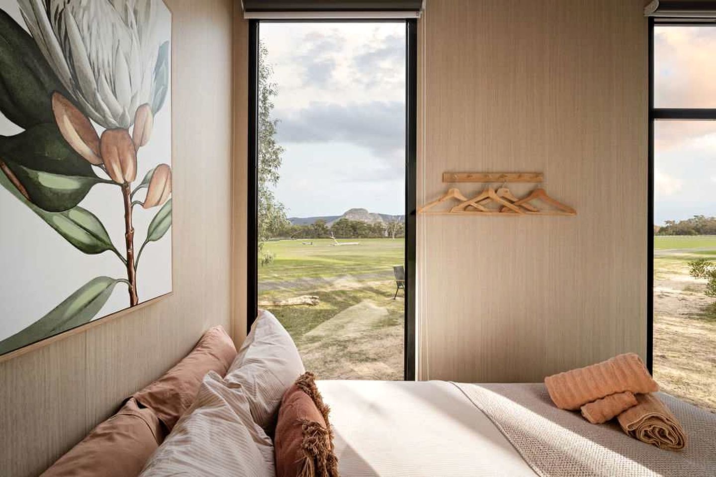 A bedroom with a large window, a painting of a plant, a bed with pillows, and a wooden hanger rack.