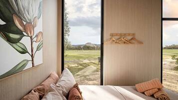 A bedroom with a large window, a painting of a plant, a bed with pillows, and a wooden hanger rack.