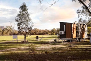 Une maison mobile dotée d’une grande fenêtre et d’une petite annexe, située dans un champ verdoyant, parsemé d’arbres, sous un ciel dégagé.
