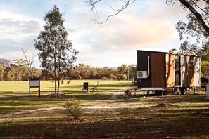 A mobile home with a large window and a small attached structure, set in a grassy field with trees and a clear sky.