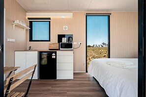 A compact bedroom with a bed, a small kitchenette, and a window with a view of a grassy landscape.