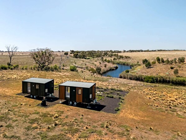 Two small cabins with metal roofs, positioned on a gravel area with a river in the background.