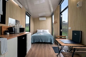 A compact room with a bed, a small kitchenette, a dining area, and a large window providing a view of the outdoors.
