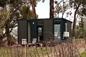 A compact, dark-colored cabin with a visible door and windows, surrounded by trees and a grassy area.