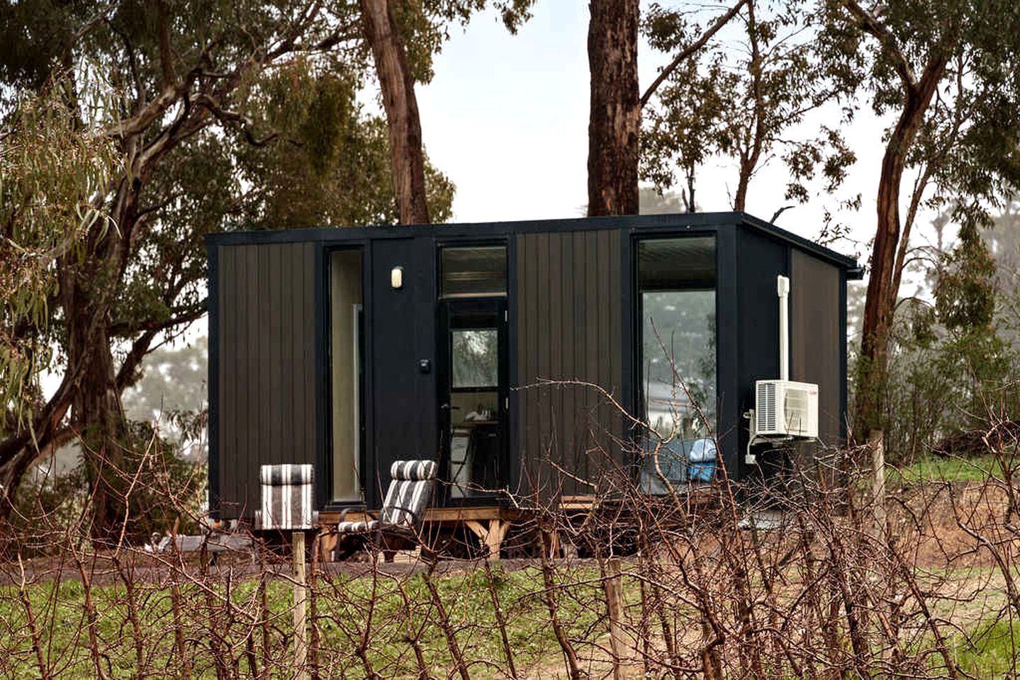 A compact, dark-colored cabin with a visible door and windows, surrounded by trees and a grassy area.