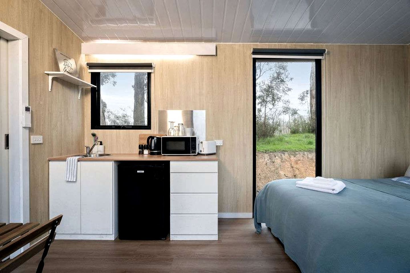 A modern bedroom with a bed, a built-in desk with a microwave, and a view of the outdoors.