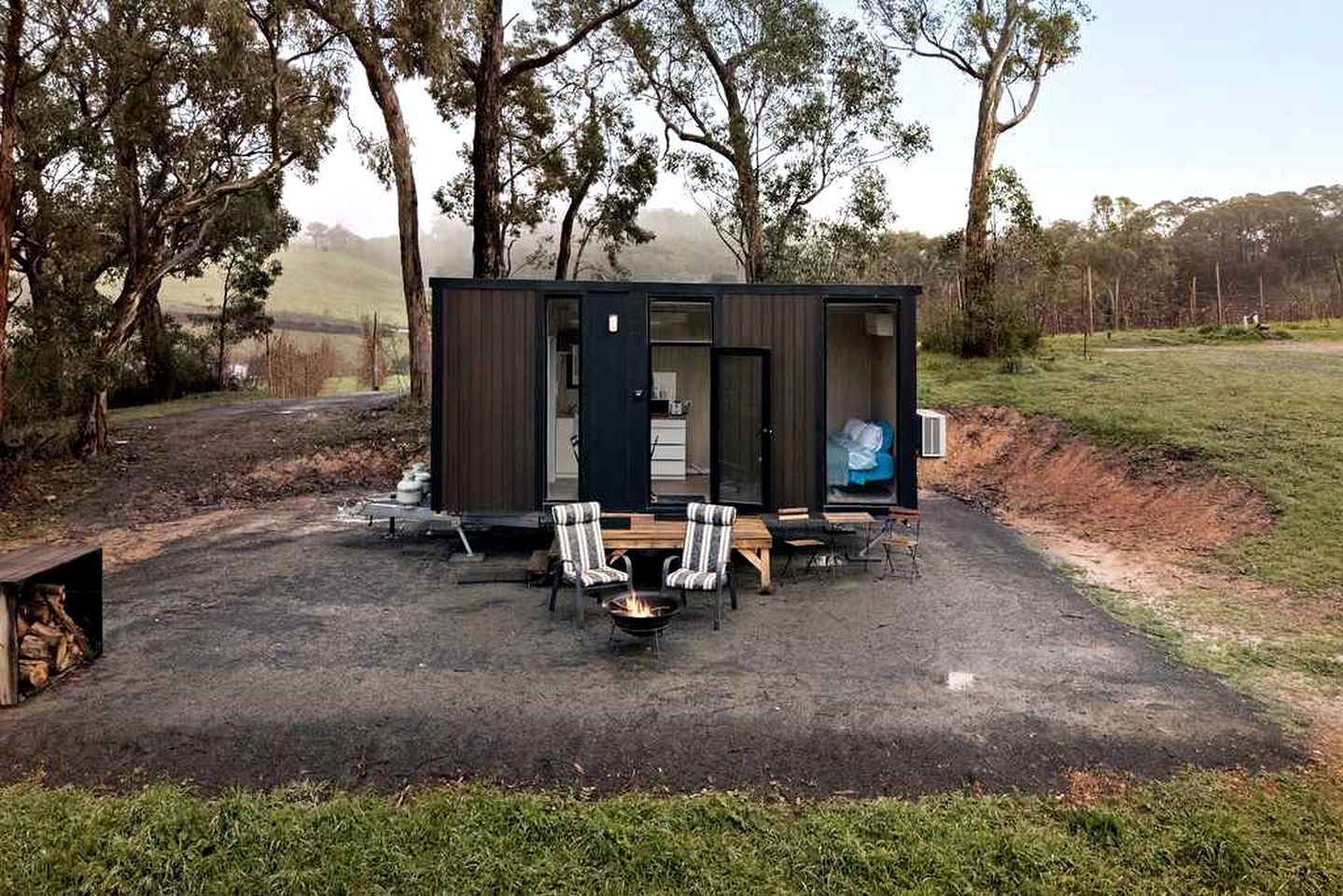 A small, dark-colored cabin with a fire pit and two chairs in front.