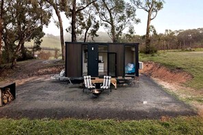 Une petite cabane de couleur sombre, avec un foyer et deux chaises devant.