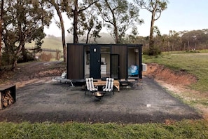 A small, dark-colored cabin with a fire pit and two chairs in front.