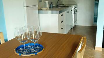 Una mesa de comedor con una bandeja azul y copas de vino, una zona de cocina con armarios blancos y un suelo de baldosas.