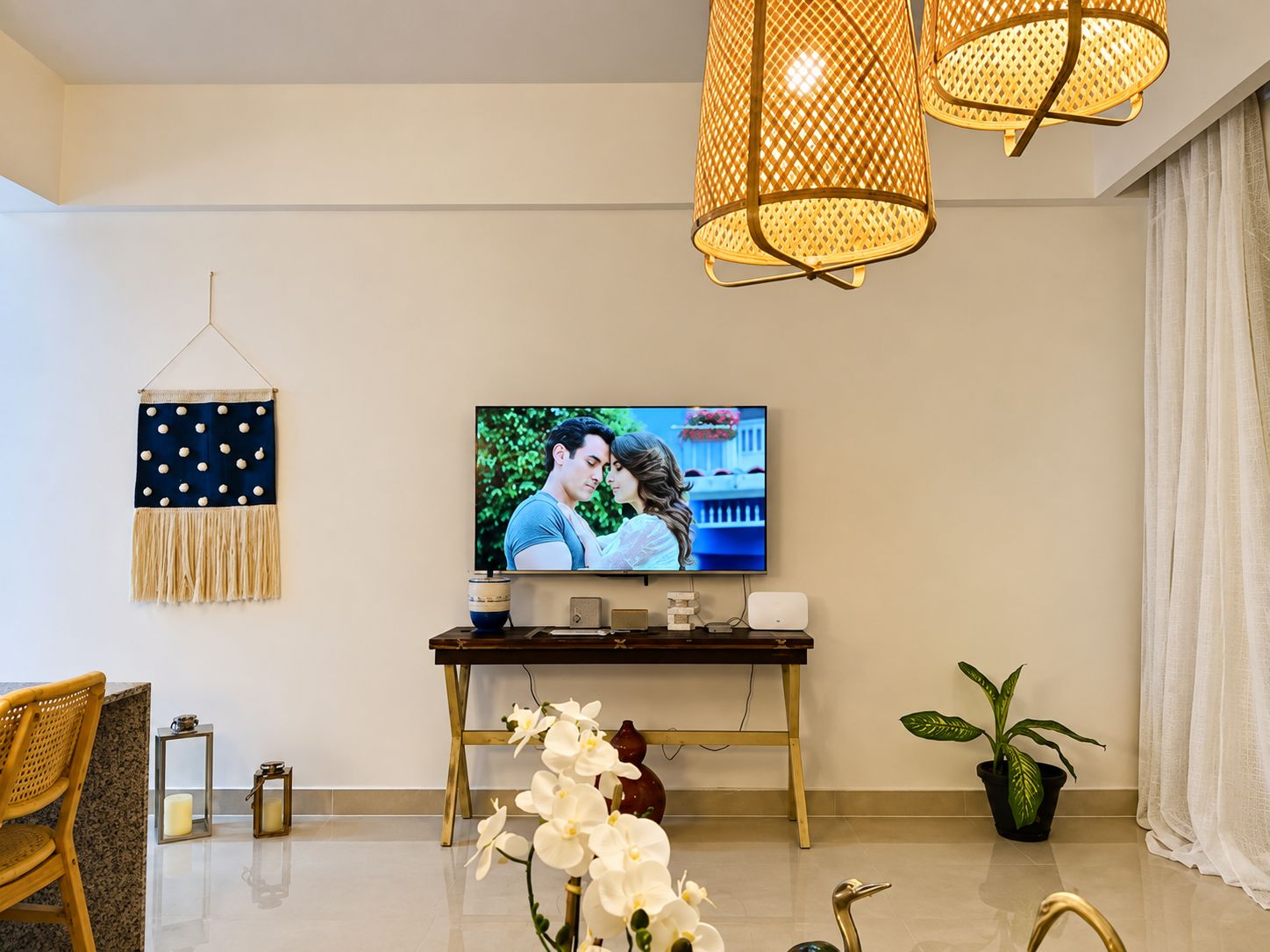Ein Wohnzimmer mit Flachbildfernseher, einem Holztisch, Wanddeko, einer Topfpflanze und einer Weidenlampe.