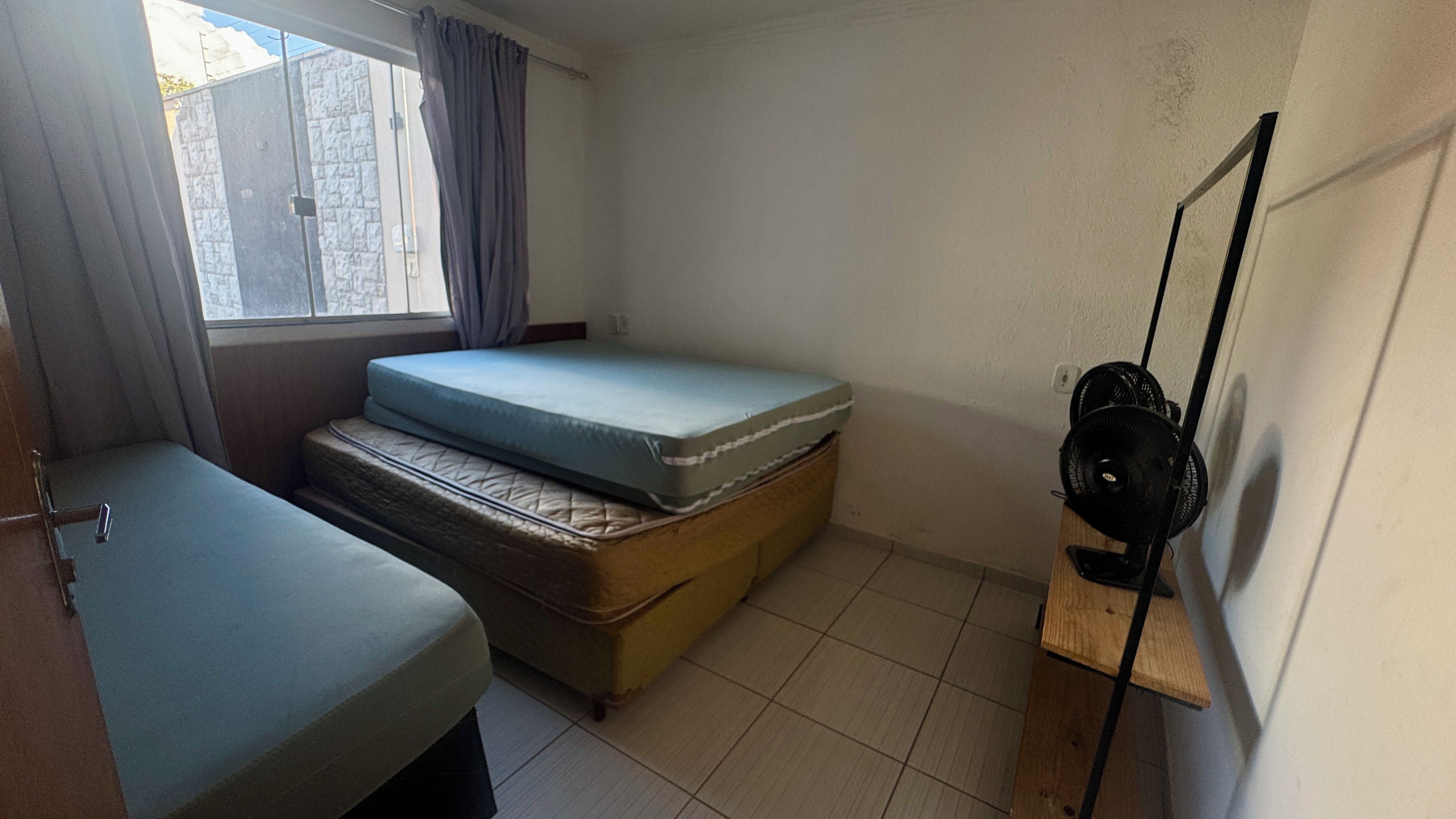 A small room with two beds, a window with curtains, a fan, and a tiled floor.