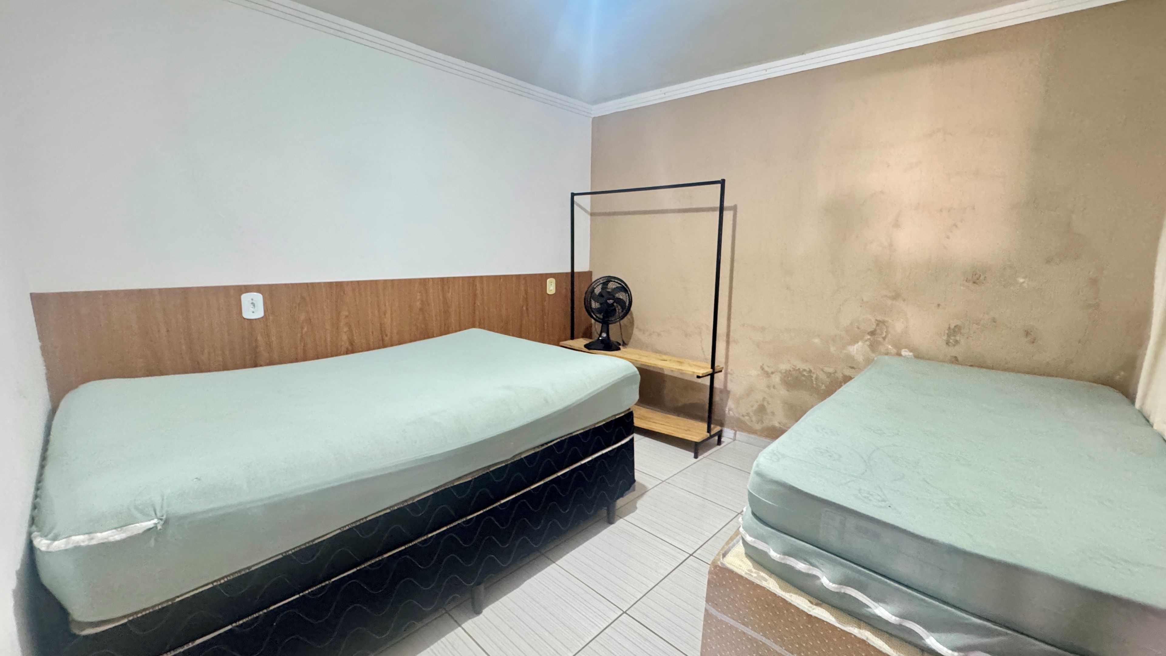 Quarto pequeno com duas camas de solteiro, uma prateleira de madeira, um ventilador e parede lisa.