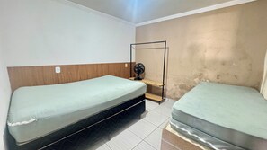 Una habitación pequeña con dos camas individuales, un estante de madera, un ventilador de pie y una pared lisa.