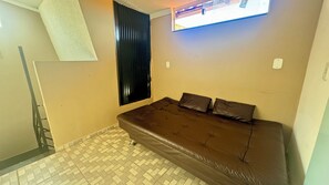 Una habitación con un sofá marrón, una ventana con persianas, piso de baldosas y una lámpara de pared.