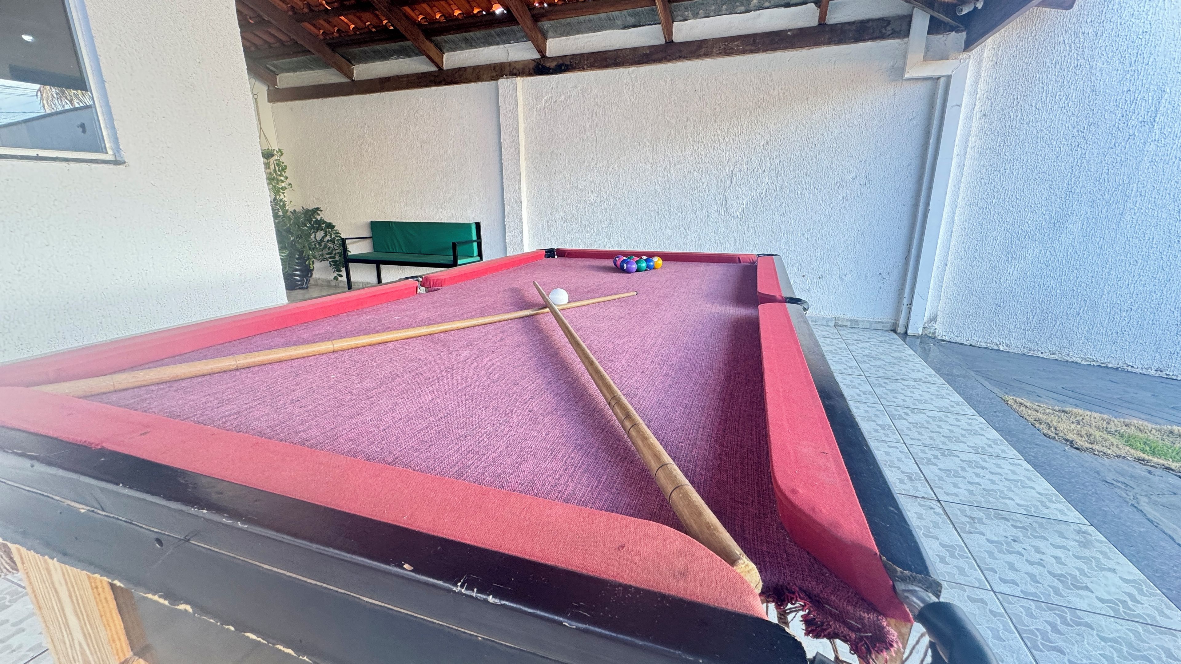Uma mesa de bilhar com superfície de feltro vermelho, rodeada por uma parede texturizada branca e um chão ladrilhado.