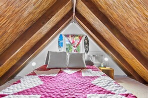 A bedroom with a sloped ceiling, a bed with a pink and white quilt, two pillows, a surfboard decoration, and a window with a view of greenery. - Peaceful Eco-Farm A-Frame in Keaau,Volcano & Beach (Keaau)