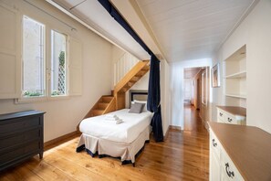 Ein Schlafzimmer mit Holzboden, einem Bett mit weißer Bettwäsche, einem Schminktisch, einer Treppe und einem Flur, der in ein weiteres Zimmer führt.