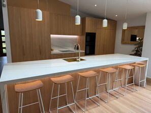 A modern kitchen with a white marble countertop, stainless steel sink, and wooden bar stools. - Spacious family getaway in the heart of Cowes, featuring a heated pool, gas-heated spa, basketball court, in-ground trampoline and outdoor kitchen with BBQ and pizza oven. Just 800m from the beach and 1km from shops and cafés. Perfect for families! (Cowes)