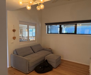 Un salon moderne avec un canapé d’angle gris, un fauteuil en forme de poire noir, un luminaire suspendu au plafond et une fenêtre donnant sur une clôture bleue.