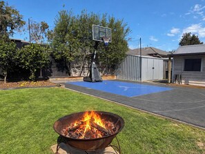 A backyard with a fire pit, a basketball hoop, and a blue rubberized play area. - Spacious family getaway in the heart of Cowes, featuring a heated pool, gas-heated spa, basketball court, in-ground trampoline and outdoor kitchen with BBQ and pizza oven. Just 800m from the beach and 1km from shops and cafés. Perfect for families! (Cowes)