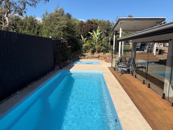 Une piscine dotée d’une terrasse en bois et de parois en verre, entourée de verdure.