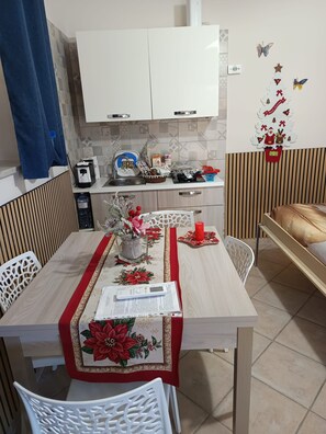 A dining area with a table set for two, a kitchen in the background, and a decorative wall with holiday ornaments. - Small house with a garden, parking space, gazebo, tables, chairs, and an outdoor kitchen. (Pagani)