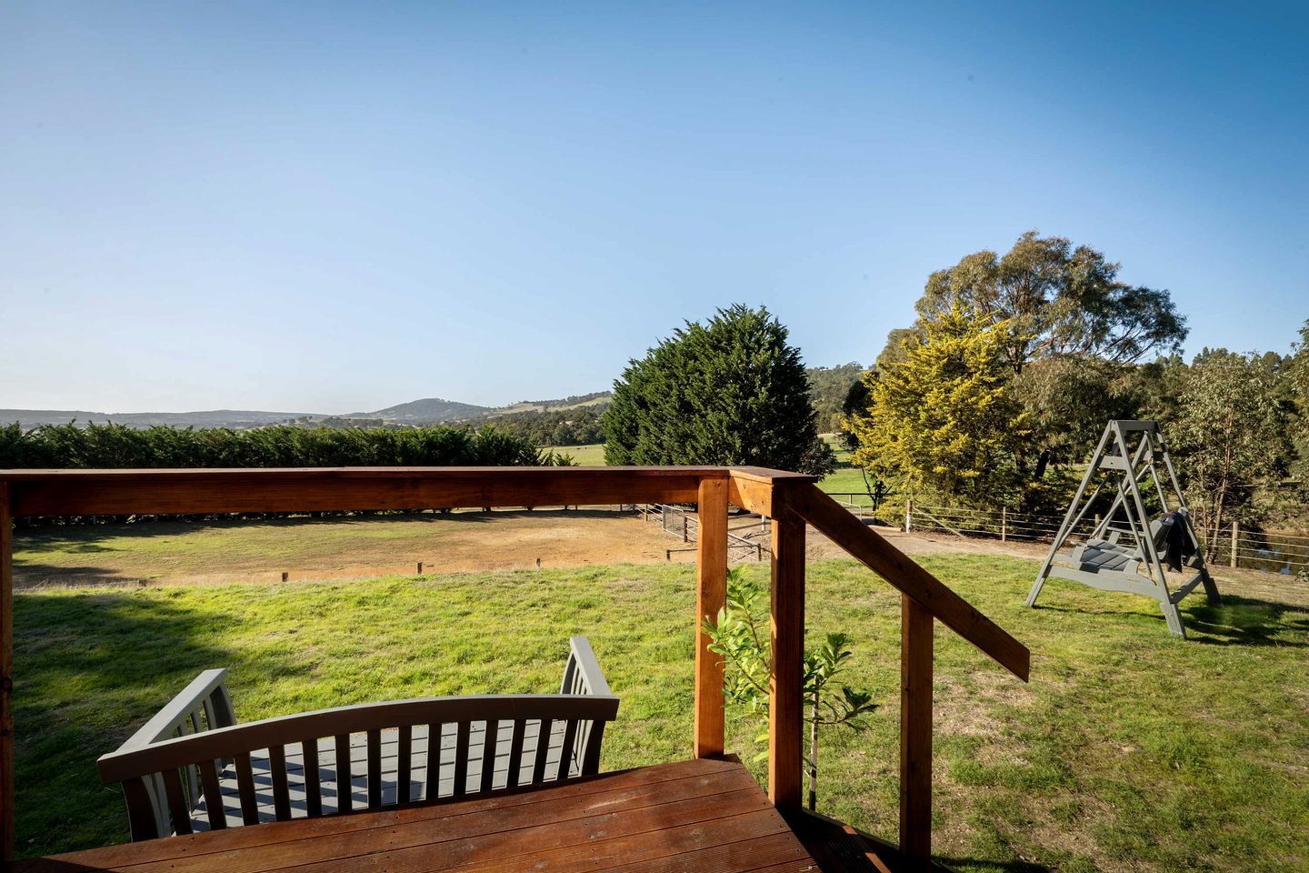 A wooden deck with a view of a grassy field and a swing set.