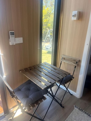 A room with a wooden bench, a cushioned seat, a wall-mounted light switch, and a window with a view of greenery.