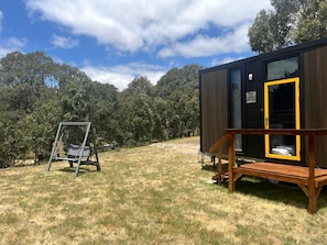 A compact wooden structure with a yellow-framed door, a swing chair, and a wooden deck.