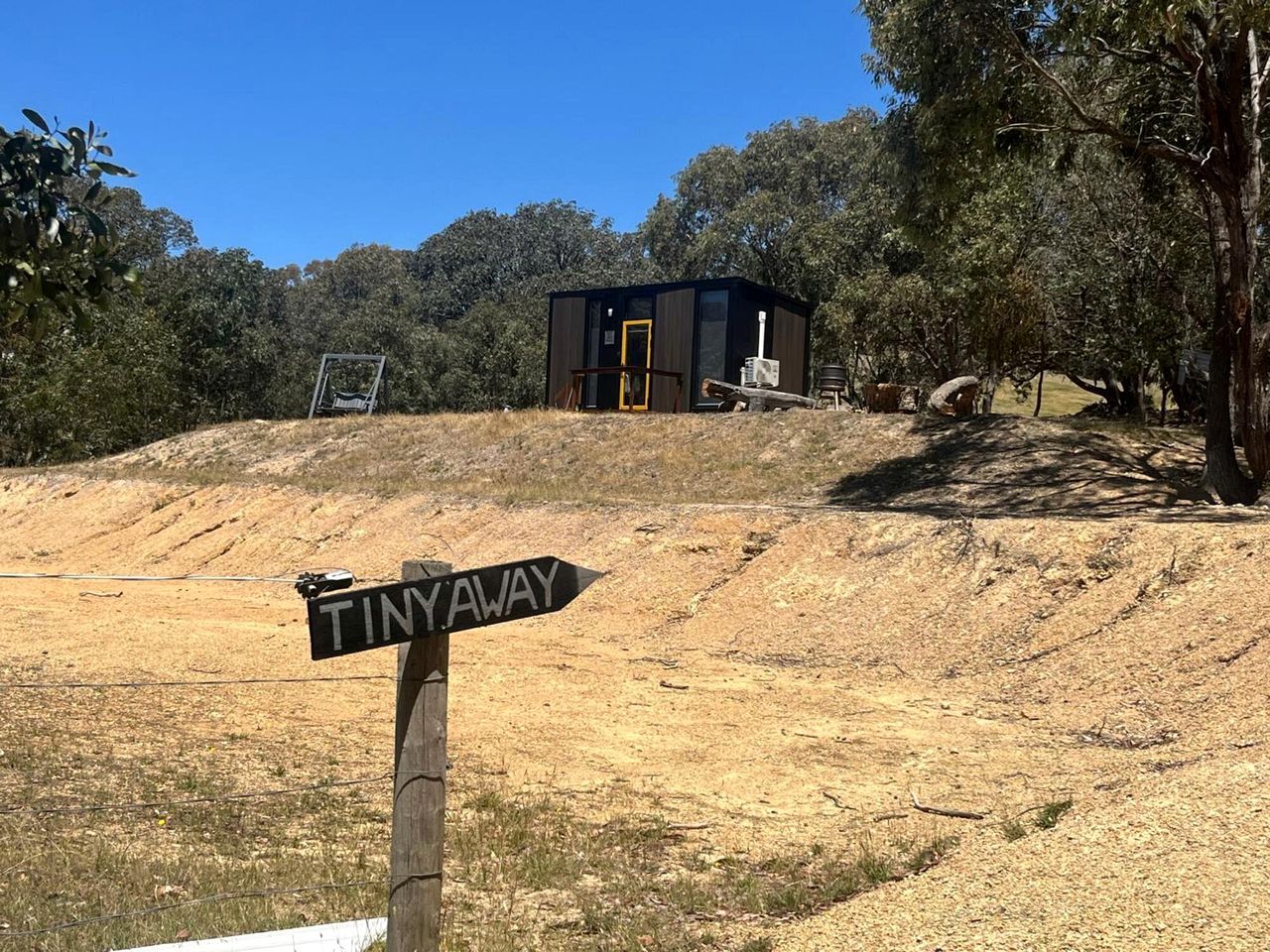 A wooden signpost with 'TINNYAWAY' written on it, pointing to a small building with a dark exterior and a yellow door.