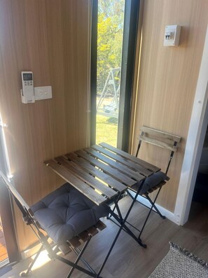 Ein Zimmer mit einer hölzernen Bank, einer gepolsterten Sitzgelegenheit, einem wandmontierten Lichtschalter und einem Fenster mit Blick auf Grünflächen.