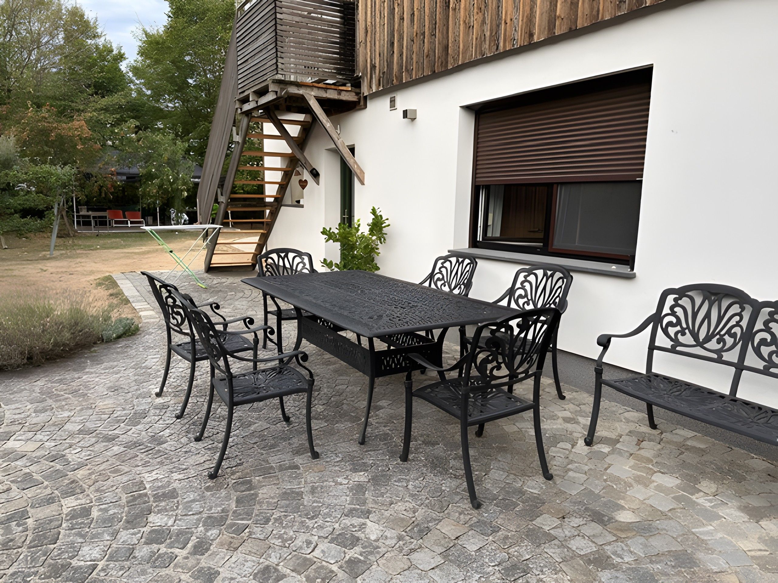 Een buitenterras met een tafel, stoelen, een houten trap en een raam met een luik.