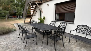 Een buitenterras met een tafel, stoelen, een houten trap en een raam met een luik.