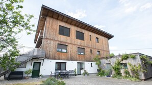 Un edificio de varios pisos con techo inclinado, fachada de madera y un patio empedrado con mobiliario exterior.