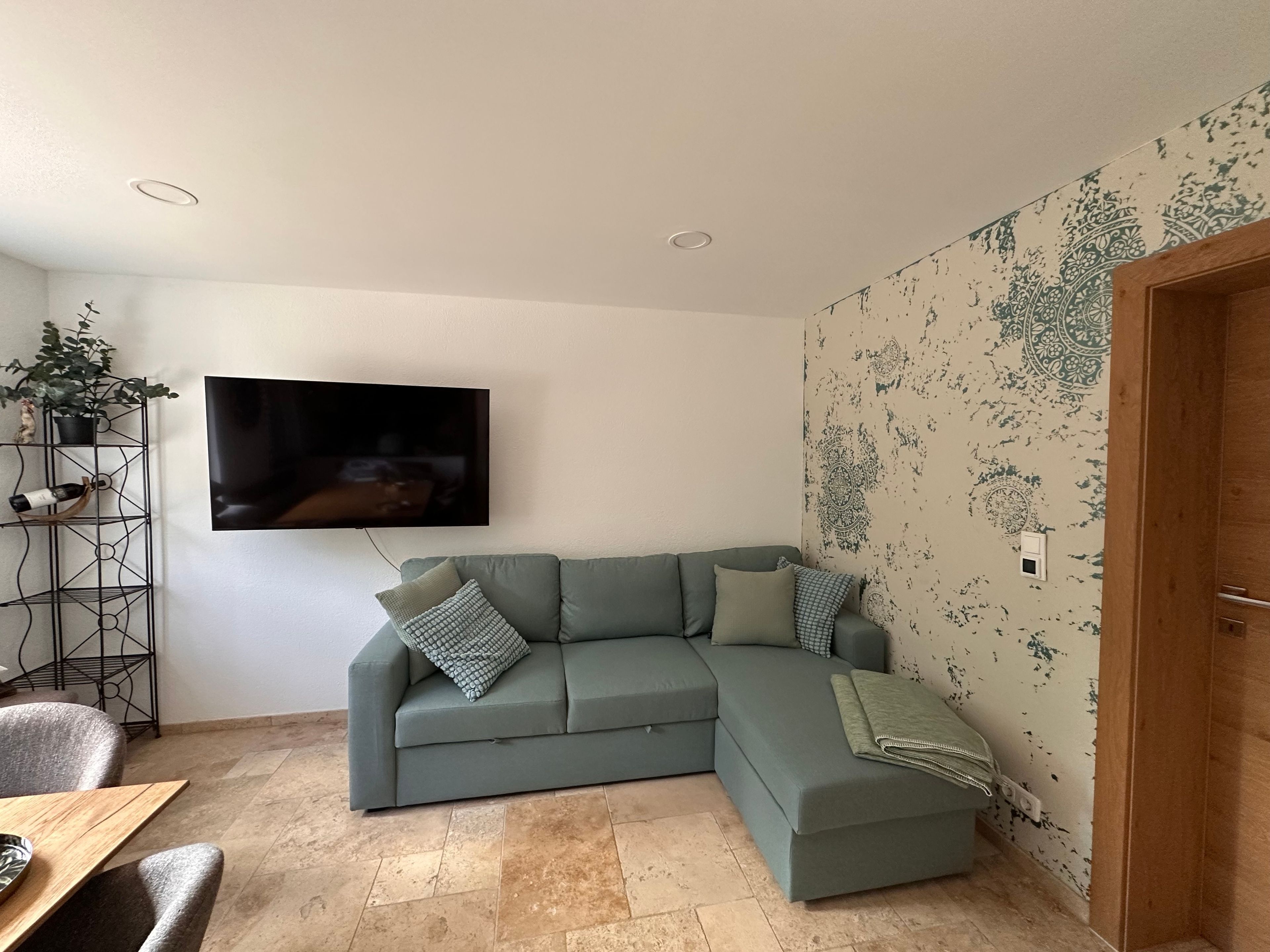 Una sala de estar con un sofá verde, un televisor montado en la pared, una planta sobre un soporte de metal y un papel tapiz estampado.
