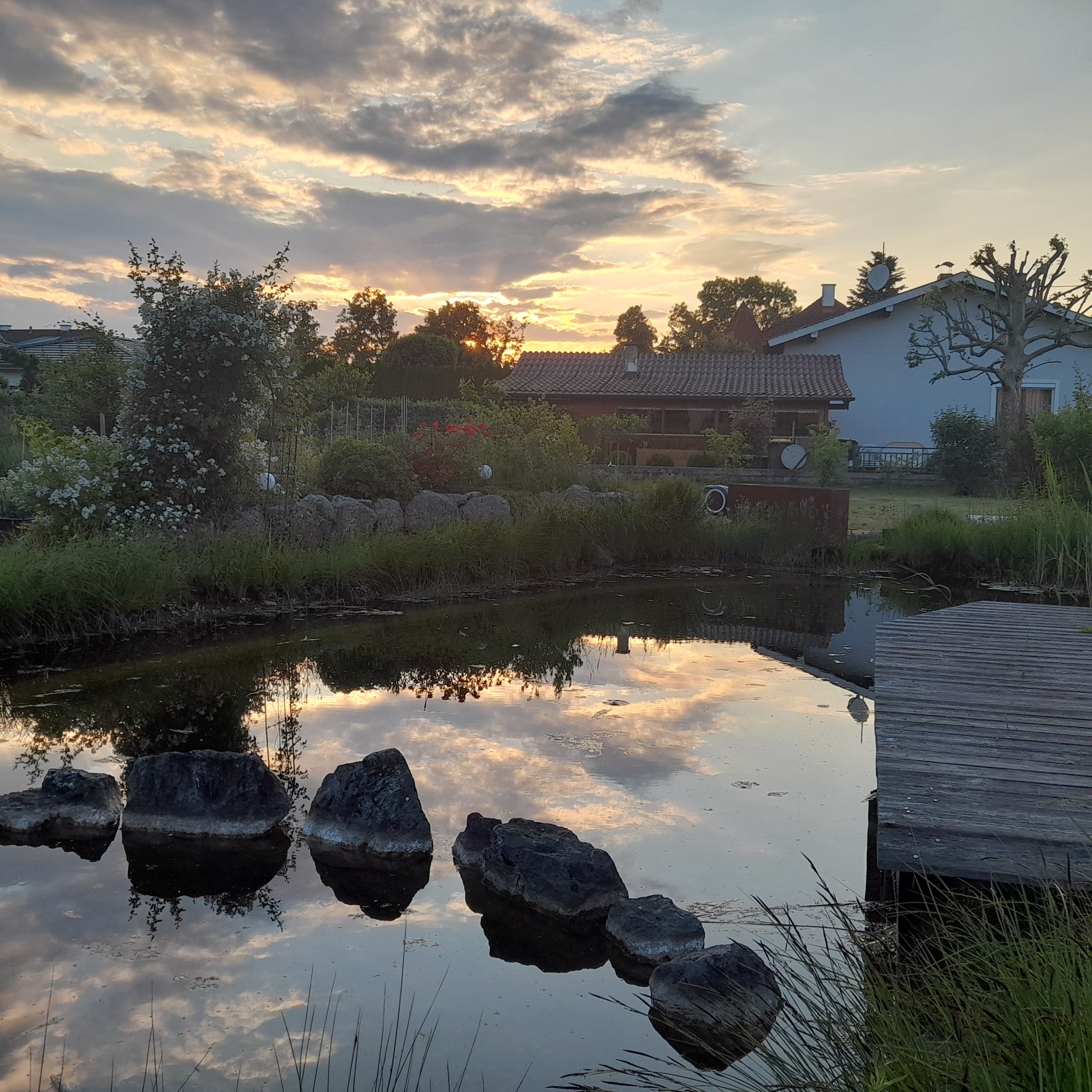 A serene riverside scene with a traditional house, a wooden bridge, and a calm water body reflecting the sky.
