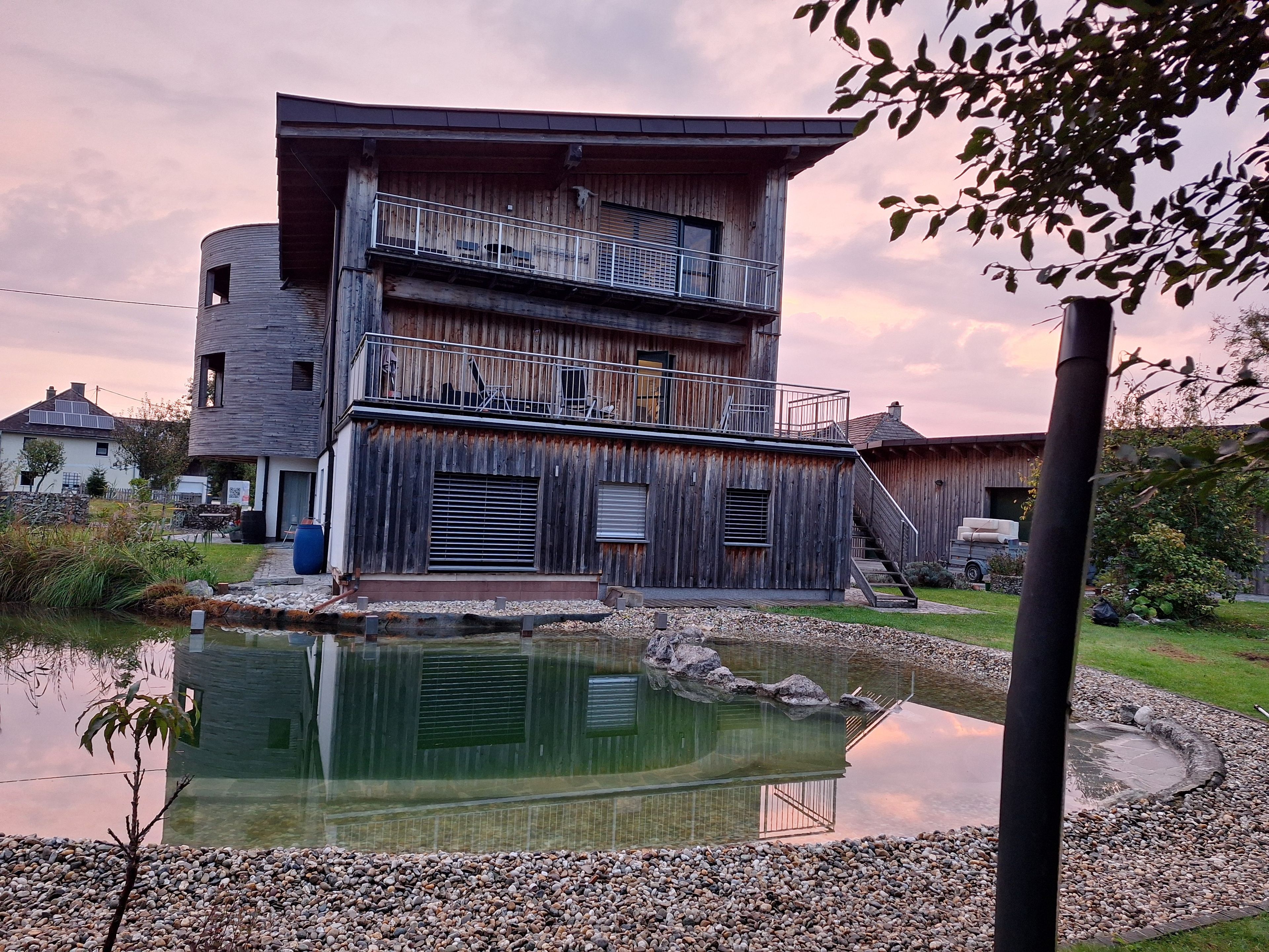 Un edificio in legno su più piani con un balcone, uno stagno e un giardino circostante.
