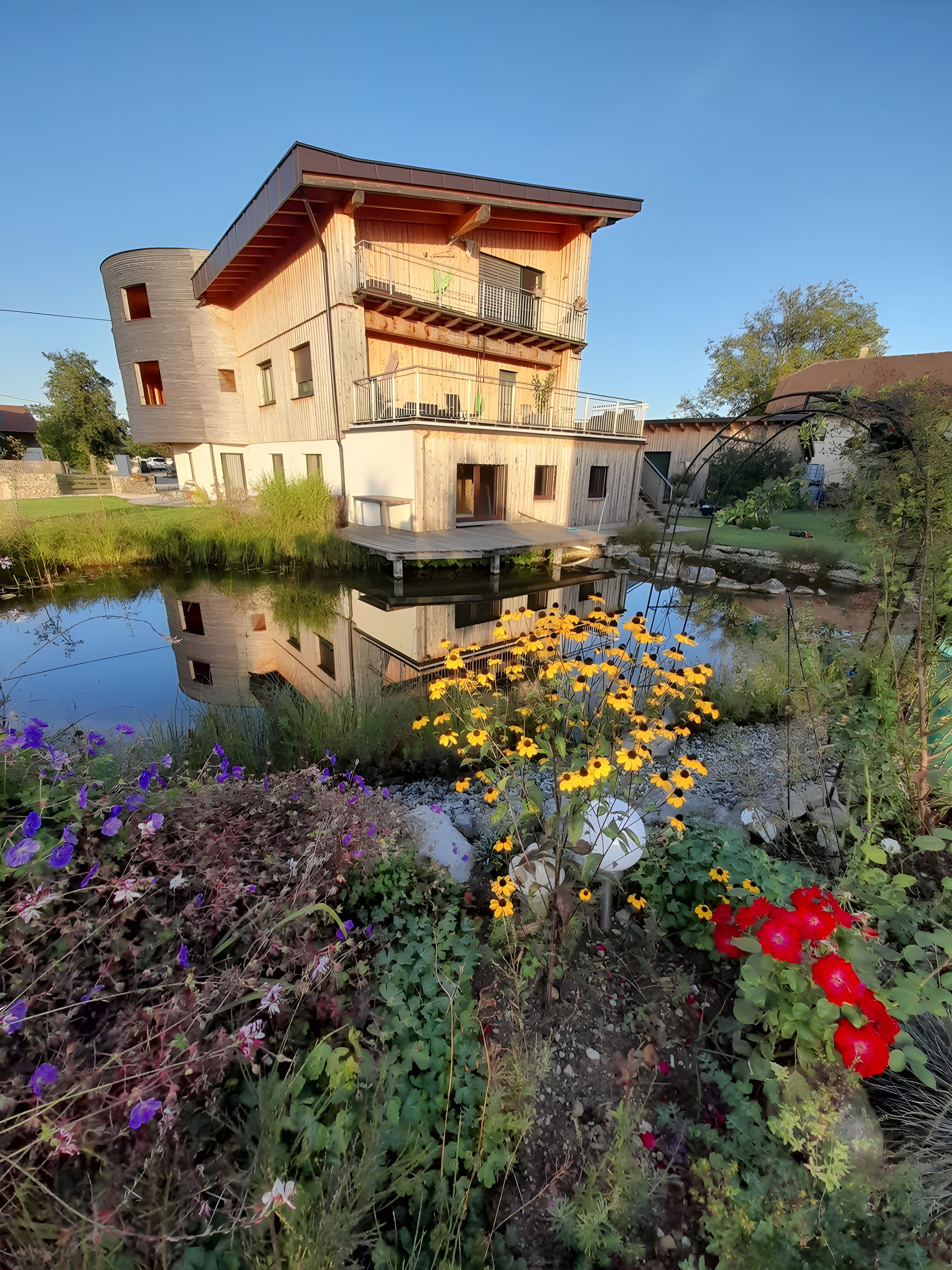 Een modern, twee verdiepingen hoog gebouw met een houten gevel en een balkon, gelegen naast een rustig waterlichaam met omliggende begroeiing.