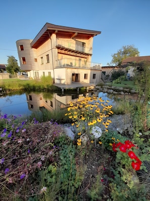 A modern two-story building with a wooden facade and a balcony, situated next to a calm water body with surrounding vegetation. - Apartment 'Gemütliche Mit Schwimmteich' with Shared Terrace, Shared Garden and Wi-Fi (Kemating)