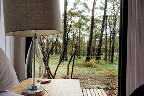 Een kamer met uitzicht op een bos, een lamp op een nachtkastje en een houten nachtkastje.