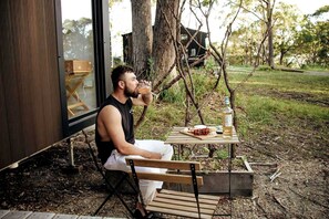 Iemand zit aan een buitentafel met een fles en eten, in een natuurlijke omgeving met bomen en een gebouw op de achtergrond.