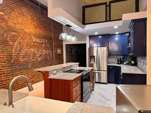 A modern kitchen with a brick wall, stainless steel appliances, and dark blue cabinets. - Stay Downtown Hot Springs! Devine's Downtown (Hot Springs)
